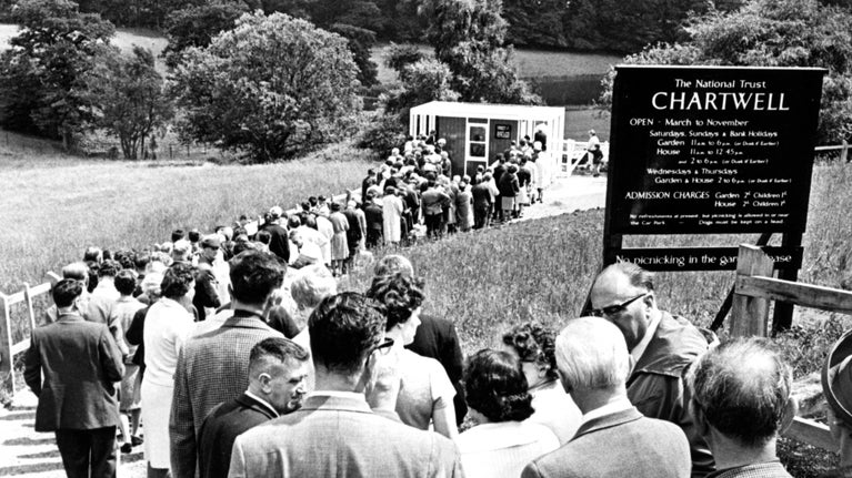 Historic image of a queue of men, women and children following a path to the ticket booth on the day The National Trust first opened Chartwell to the public 22 June 1966. A sign with opening times and prices is in the grass beside the queue of people.